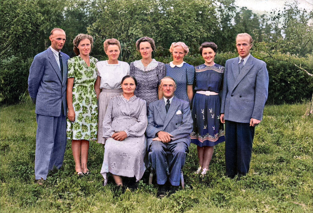 A vintage color photo of seven women and two men standing behind an older seated couple, all dressed formally, posing outdoors on green grass with trees in the background.