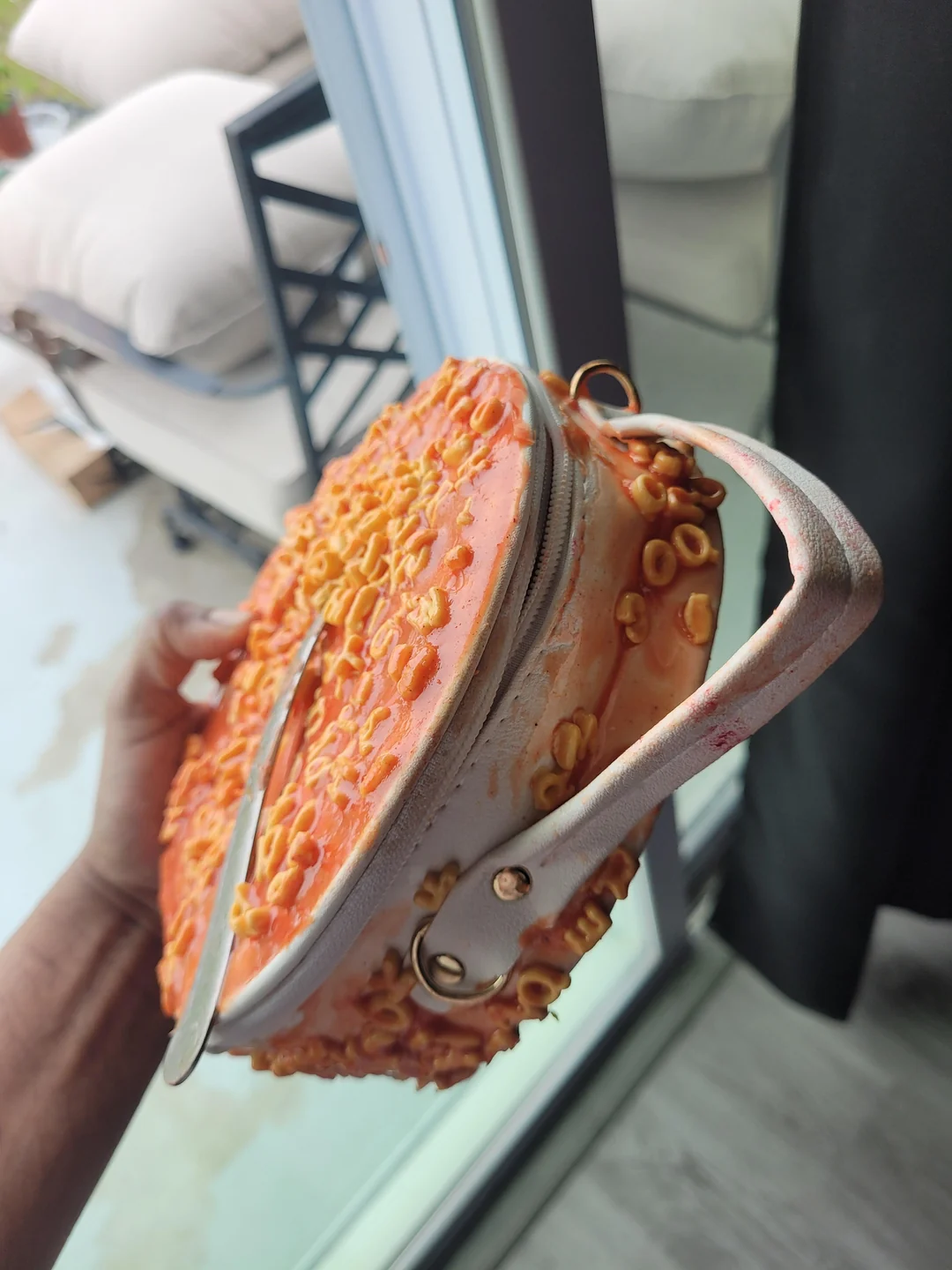 A hand holds a round purse designed to look like a dish of cheesy pasta, with orange pasta pieces and melted cheese detailing on the surface and zipper. A fork is tucked under the strap.