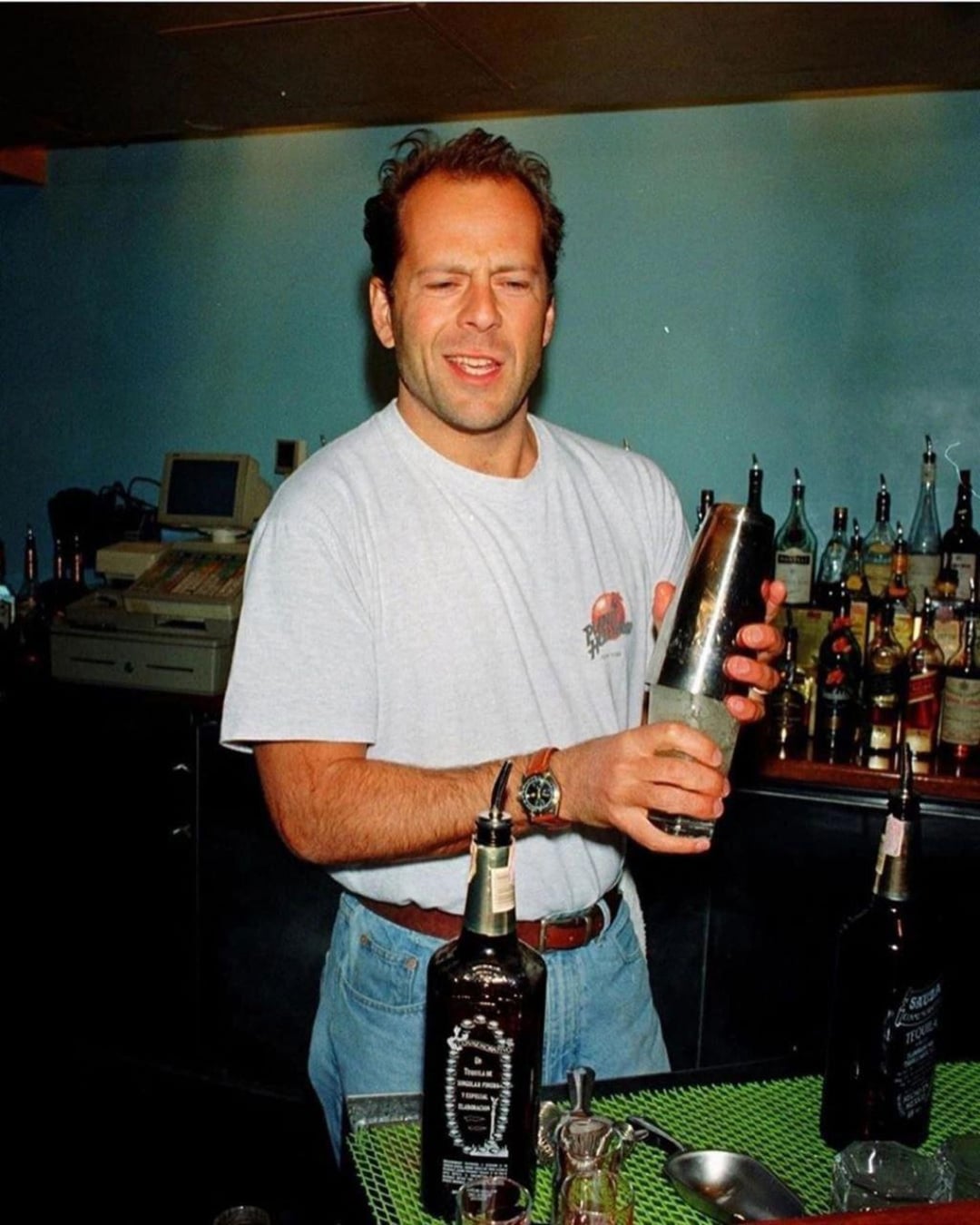 A man in a white T-shirt and jeans stands behind a bar, holding a cocktail shaker with bottles and bar tools around him. He appears to be mixing a drink and is making a facial expression as he works.