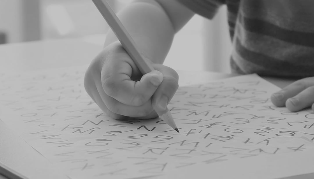 A child’s hand holds a pencil, writing letters and symbols on a sheet of paper. The child wears a striped shirt. The image is in black and white.