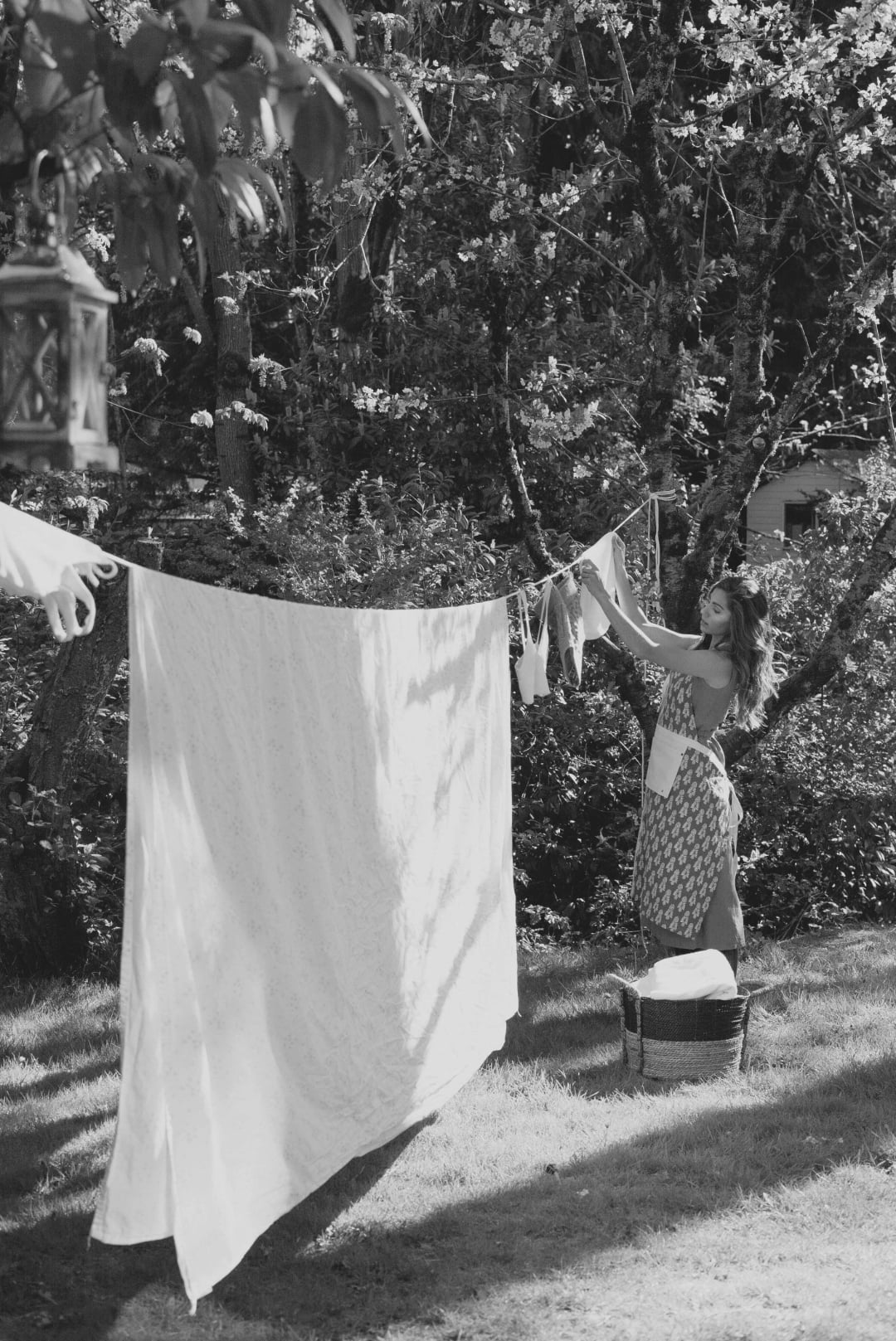 A woman hangs laundry on a clothesline in a sunlit garden, surrounded by trees. A patterned cloth is tied around her waist, and a laundry basket sits on the grass nearby. The scene has a peaceful, vintage feel.