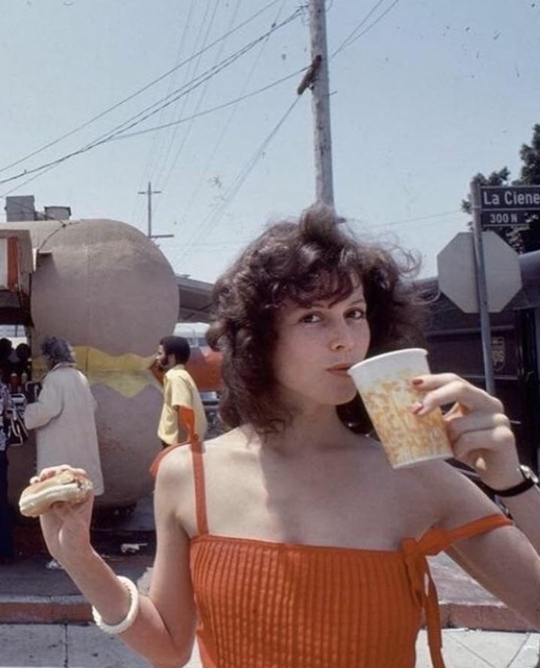A young woman in a red dress sips from a yellow drink cup and holds a hot dog. Behind her, people walk near a giant hot dog statue on a sunny street corner with visible power lines and street signs.