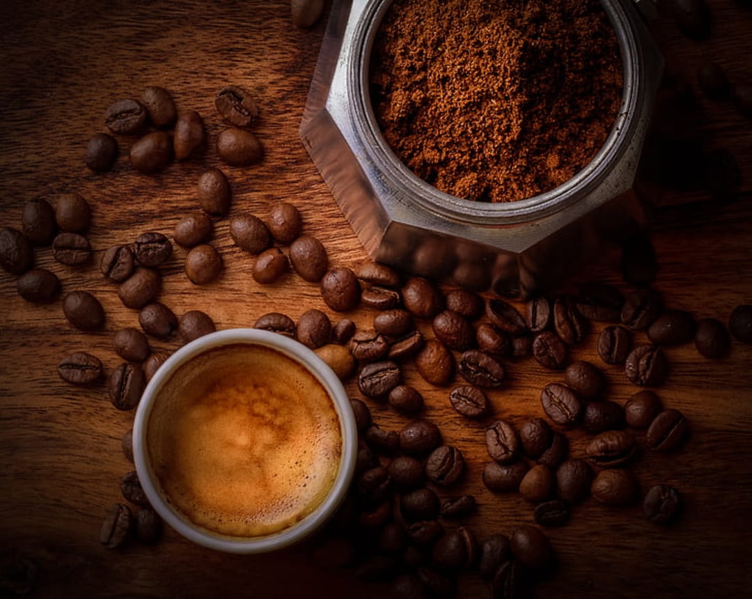 A cup of espresso sits on a wooden surface next to scattered coffee beans and an open moka pot filled with ground coffee.