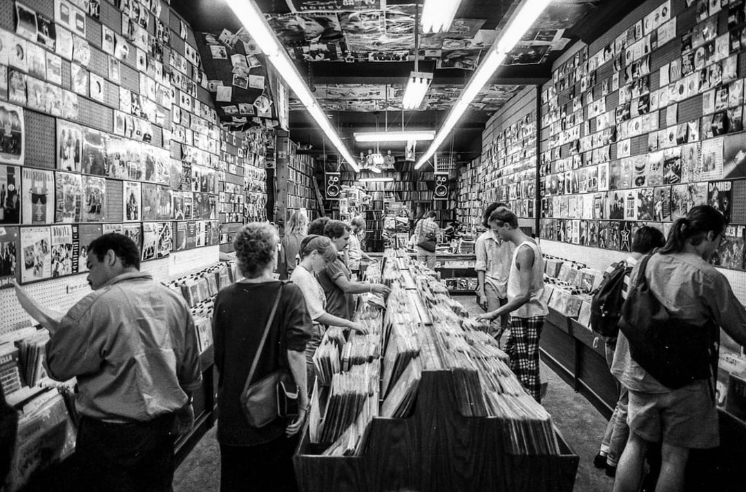 Black and white photo of people browsing records in a crowded music store. Album covers fill the walls and bins of vinyl records line the center. The scene feels busy and nostalgic.