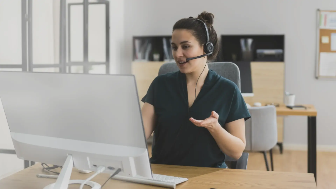 A woman wearing a headset sits at a desk, speaking and gesturing while looking at a computer monitor in a modern office setting.