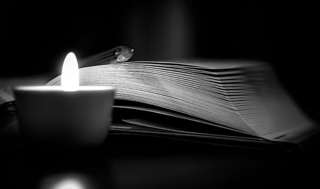 A lit candle sits in front of an open book, casting a soft glow on the pages in an otherwise dark environment. The scene is black and white, emphasizing the contrast between light and shadow.