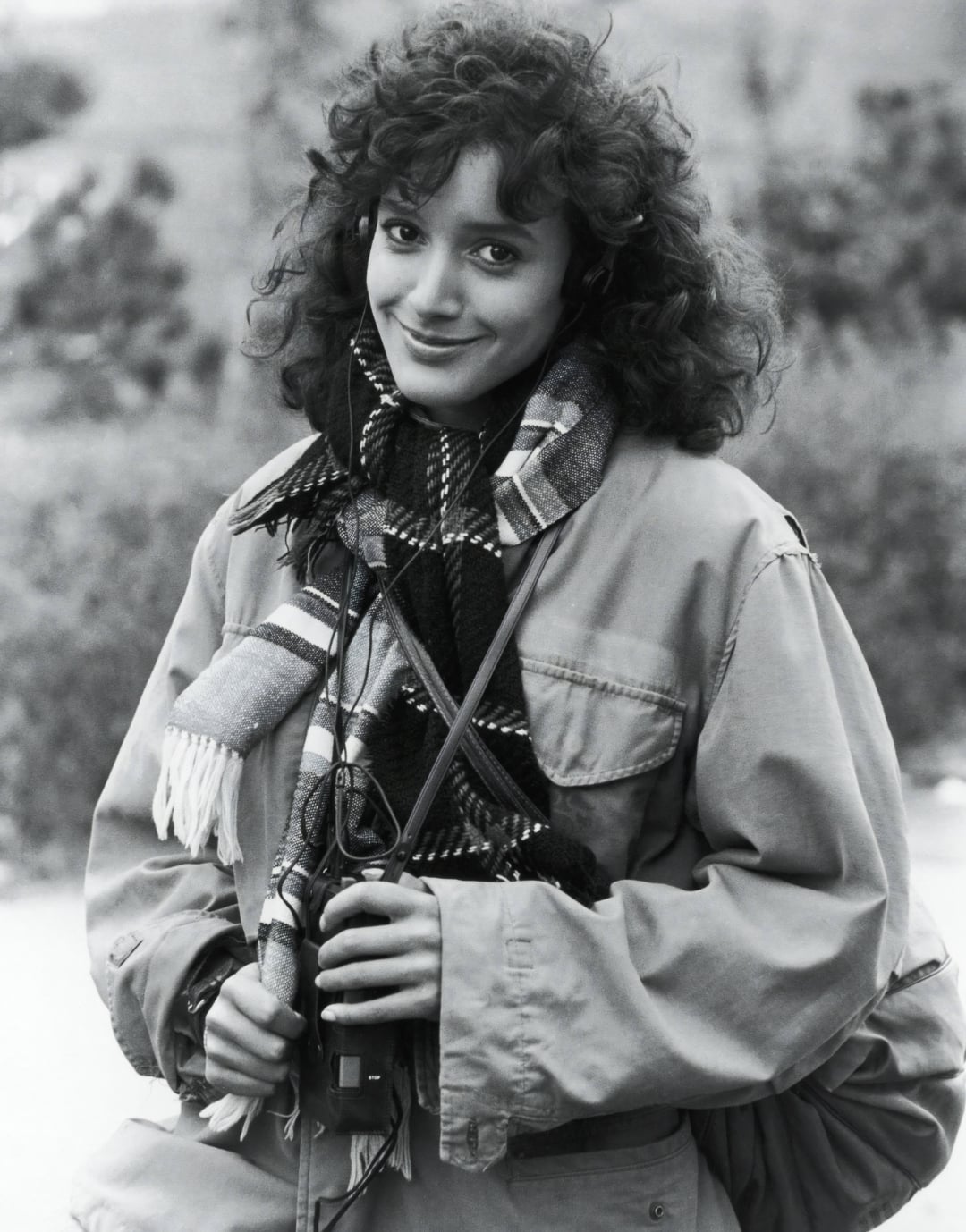 A young woman with curly hair smiles at the camera, wearing a large jacket and patterned scarf. She holds binoculars and stands outdoors with a blurred background of trees. The photo is in black and white.