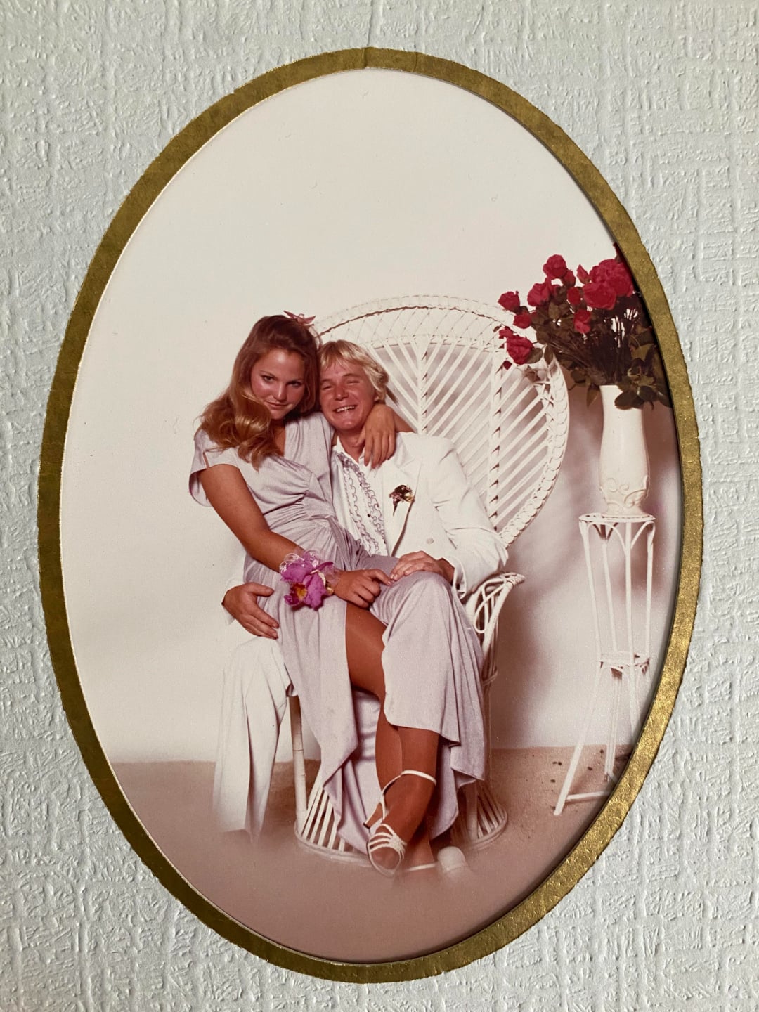 Two women dressed in light-colored formal wear pose together; one sits on the other's lap in a white wicker chair next to a vase of red roses, all within a gold-framed oval photo against a textured wall.