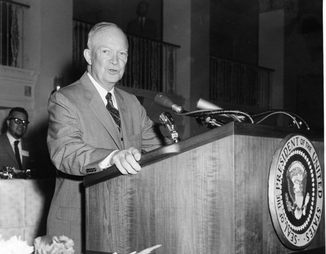 An older man in a suit speaks at a podium with the Seal of the President of the United States. He stands indoors, and a microphone and documents are on the podium. Another man is visible in the background.