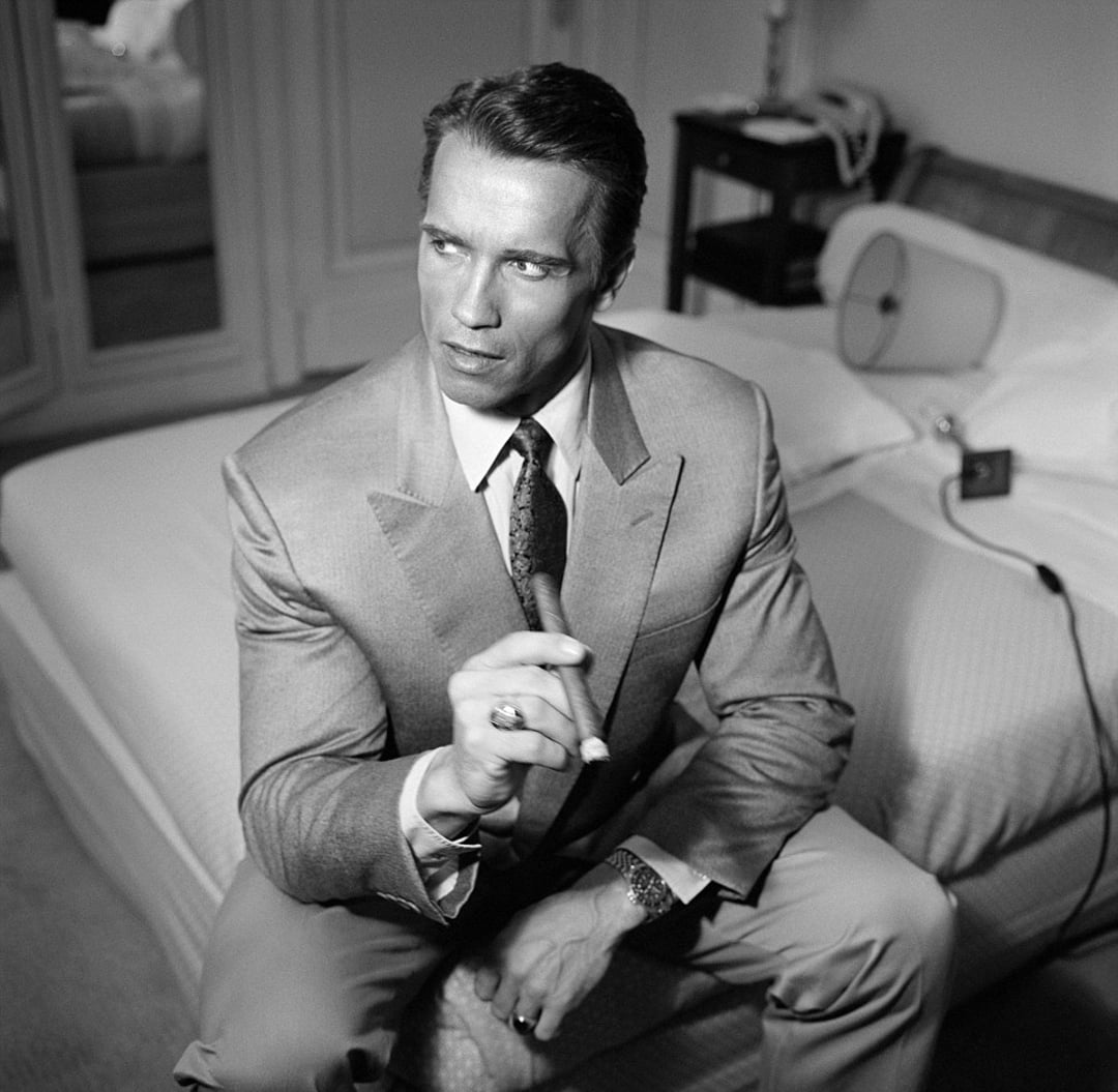 A man in a suit sits on the edge of a bed, holding a cigar and looking to the side. The room has a classic decor, with a bedside table, lamp, and telephone visible in the background. The image is in black and white.