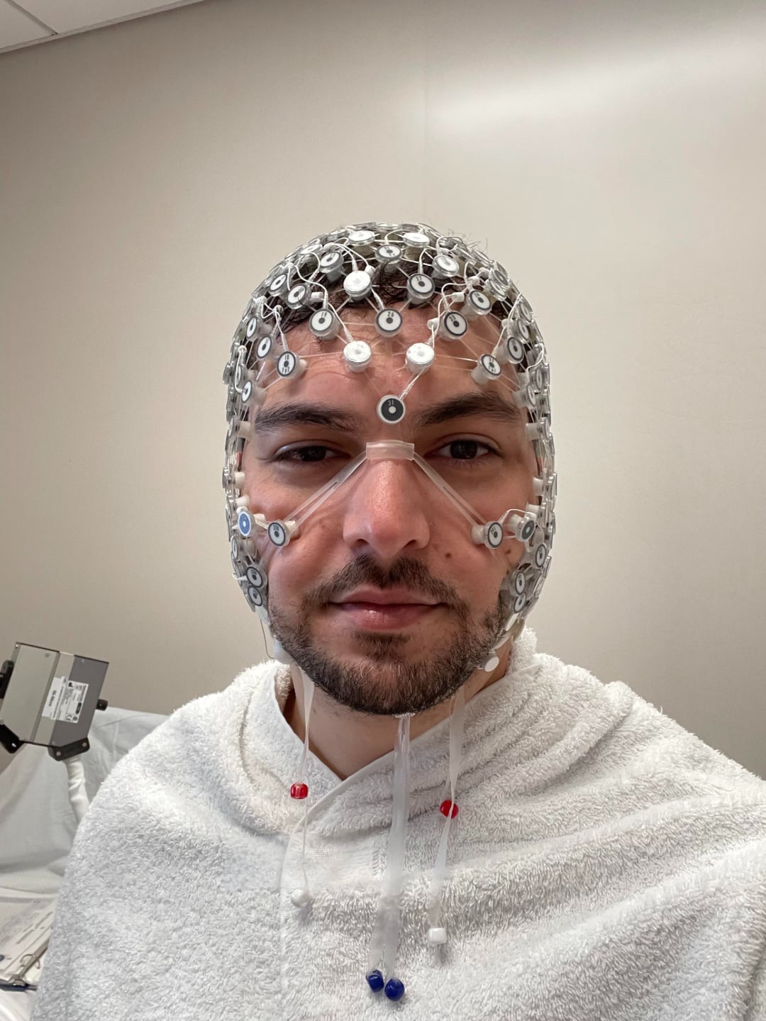 A person wearing a medical cap covered in electrodes and wires, likely for an EEG test, is in a clinical setting and dressed in a white robe or gown.