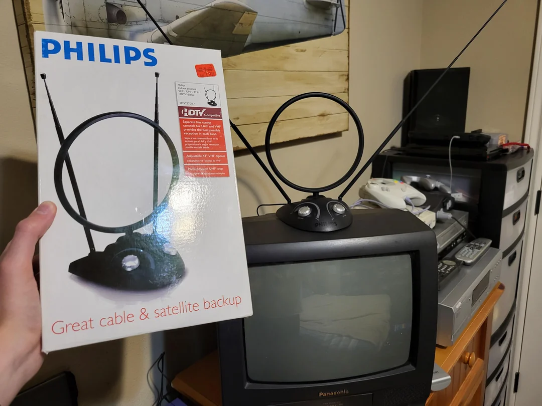 A hand holds a Philips TV antenna box in front of an old TV with the same antenna on top. The TV is part of a retro setup with a VCR, game console controllers, and drawers in the background.