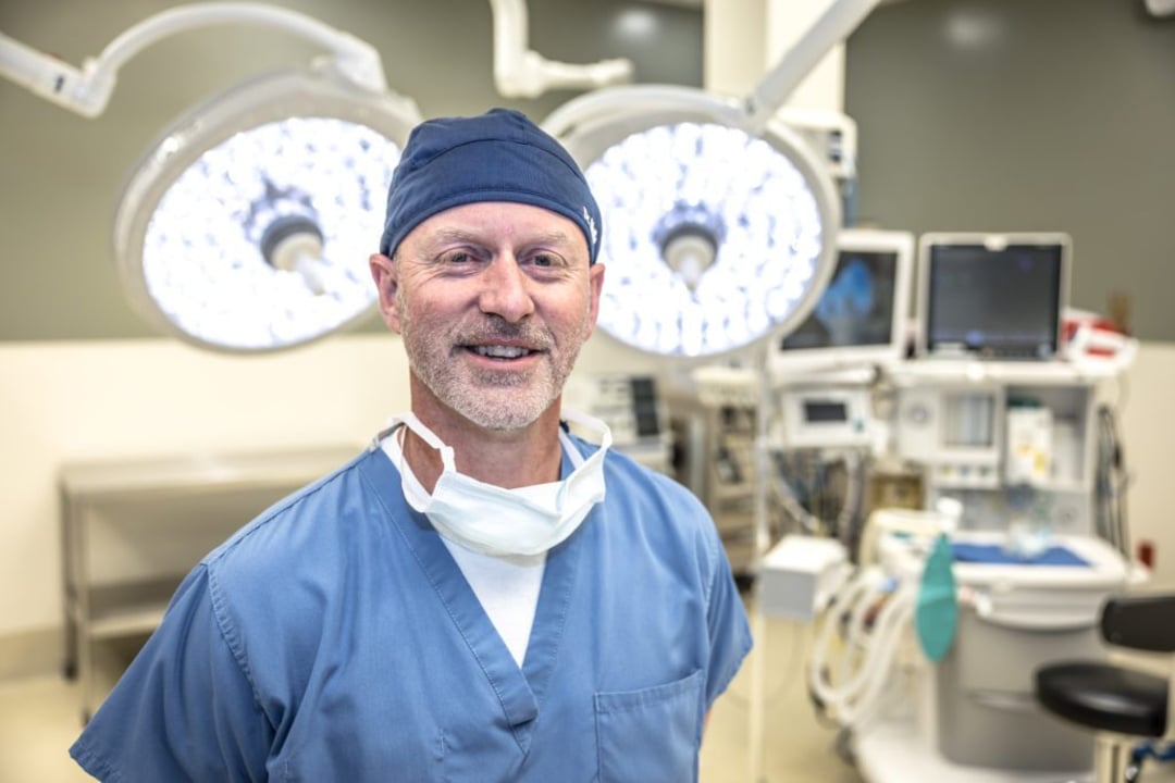 A smiling male surgeon in blue scrubs, a cap, and a mask around his neck stands in a well-lit operating room with surgical lights and medical equipment in the background.