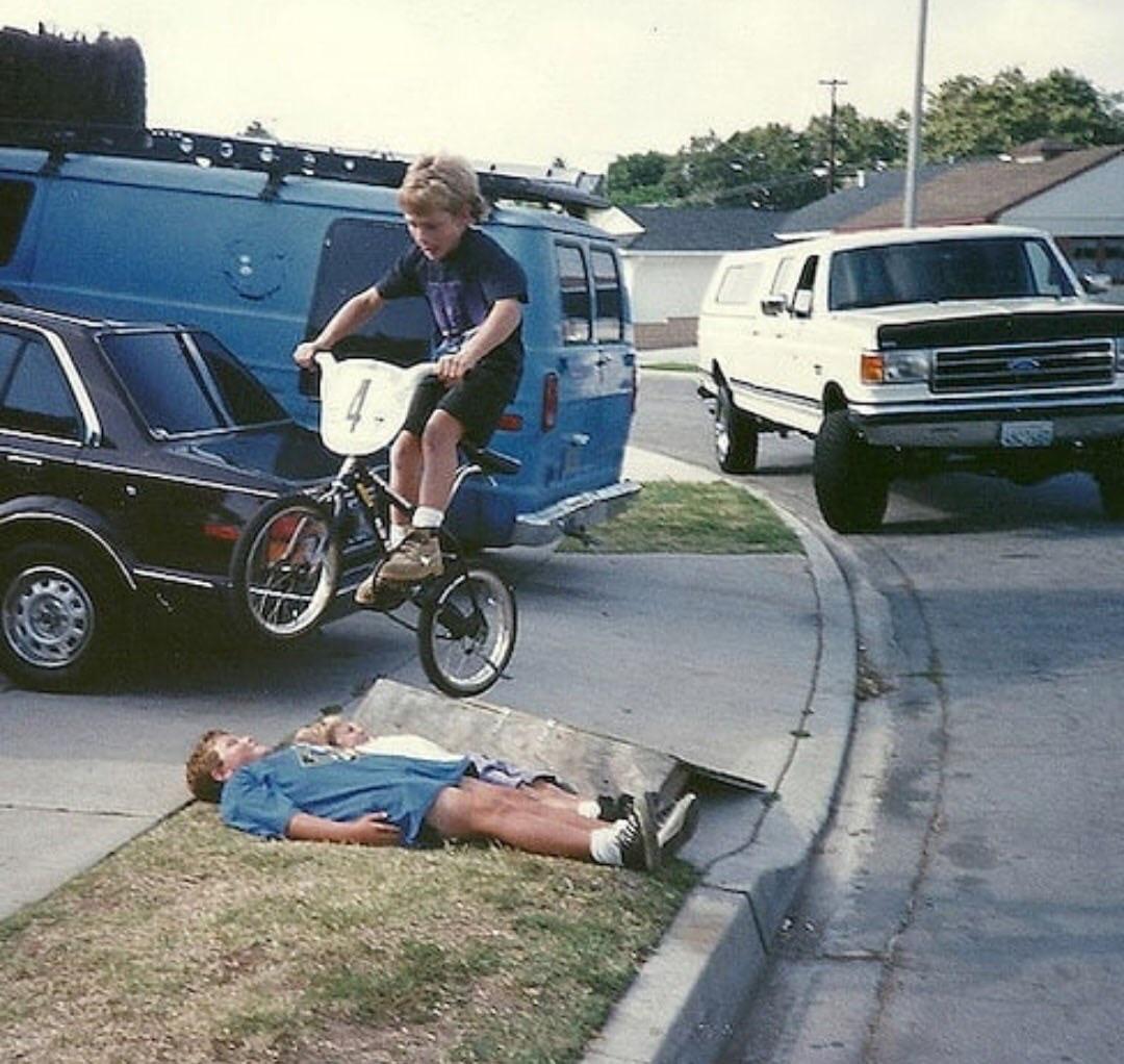 A boy on a BMX bike jumps over another boy lying on the sidewalk in a suburban neighborhood, with parked cars and houses in the background.