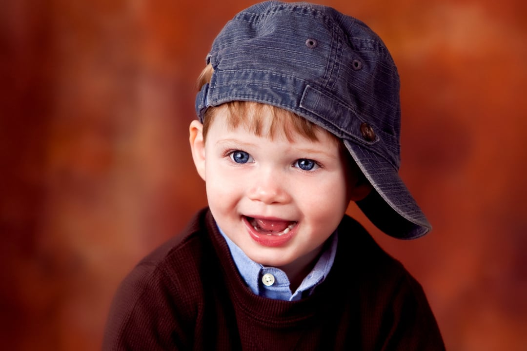 A young child with blue eyes, wearing a brown sweater and a blue cap turned sideways, smiles in front of a warm, blurred orange-brown background.