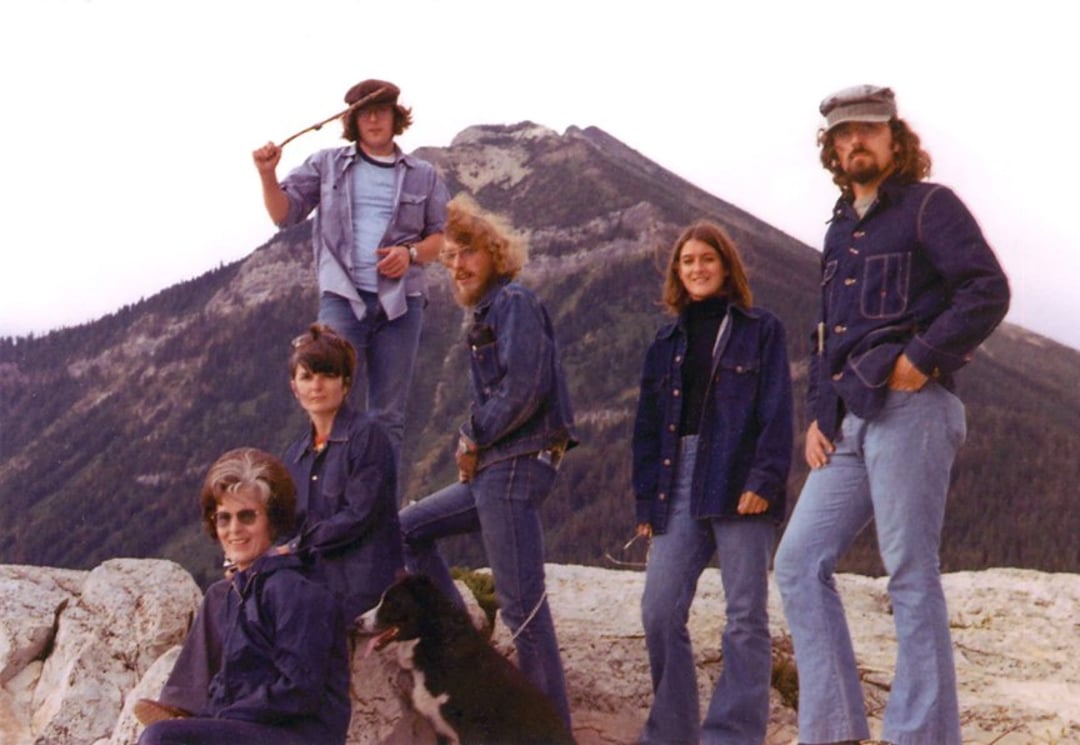 Six people in denim outfits pose outdoors on rocks with a dog. There are mountains and trees in the background. One person is standing with a raised arm, and everyone appears relaxed and happy.