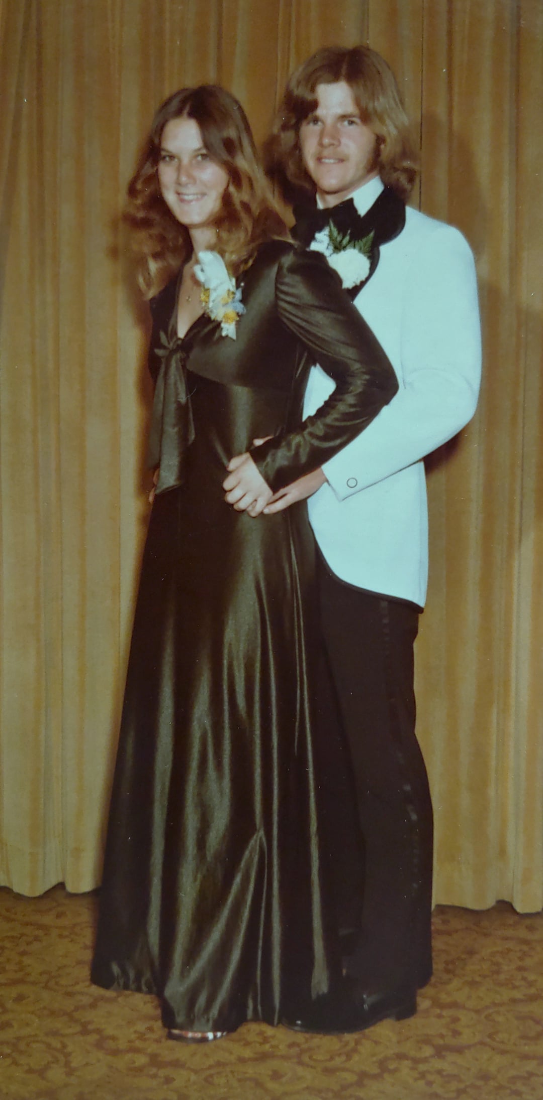 A young couple dressed in formal attire stands smiling in front of gold curtains; the woman wears a long dark gown with a corsage, and the man wears a light jacket with a boutonniere.