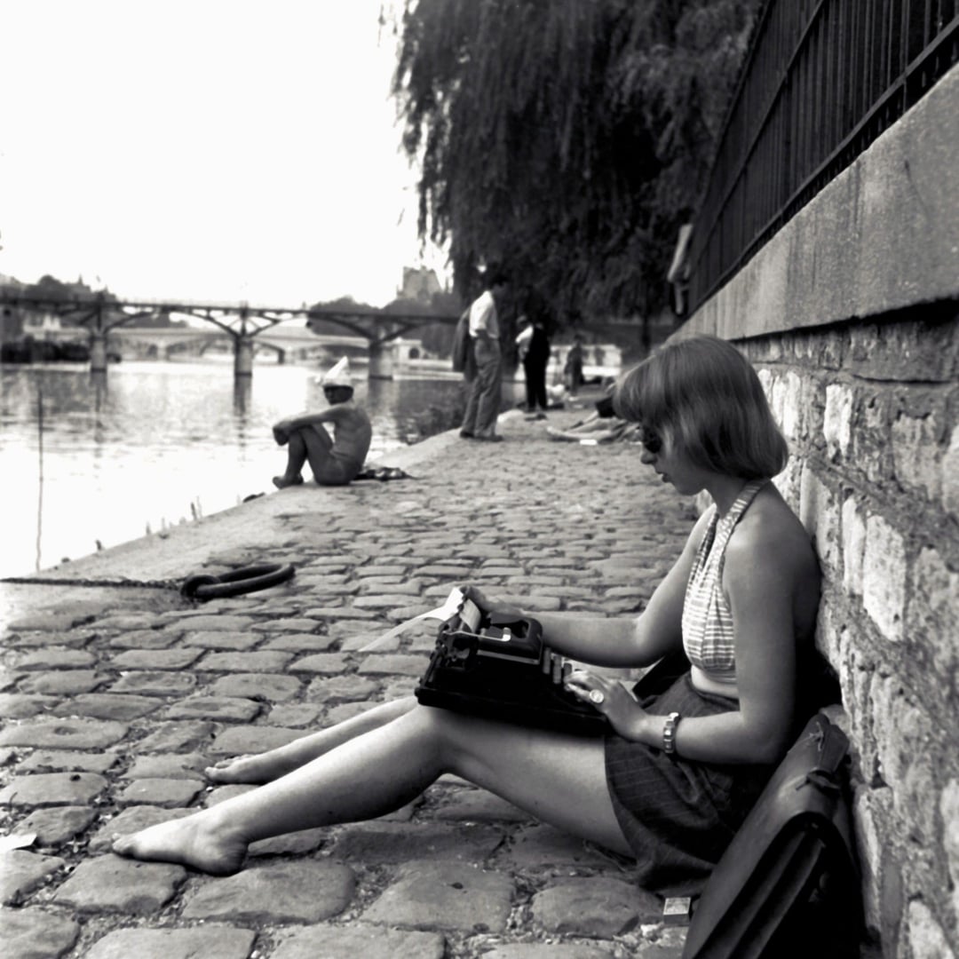 A woman sits barefoot on a cobblestone path by a river, typing on a typewriter. Other people relax nearby near the water. A bridge and trees are visible in the background.