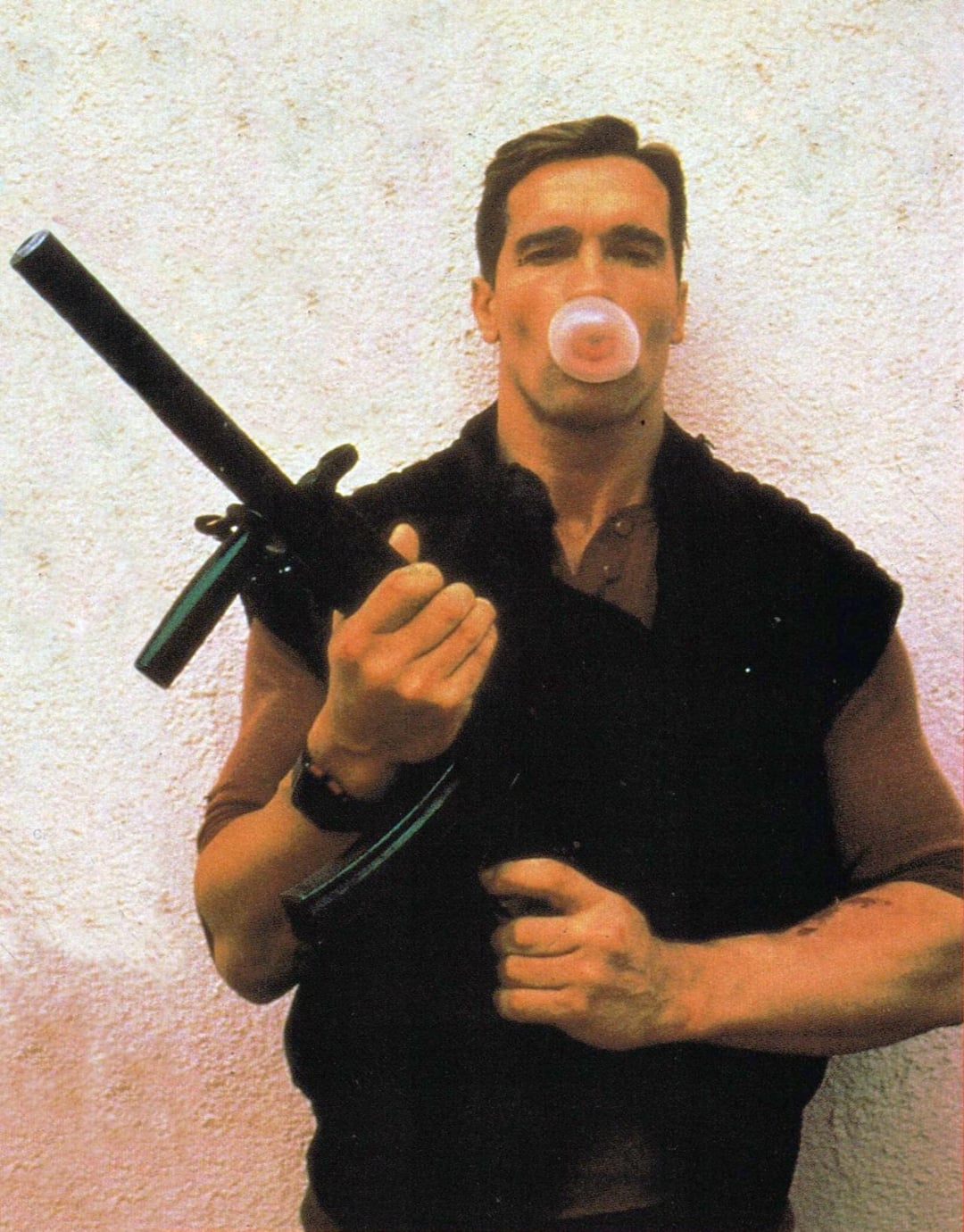 A muscular man stands against a textured wall, blowing a bubble with chewing gum while holding a large firearm in his right hand and looking straight ahead.