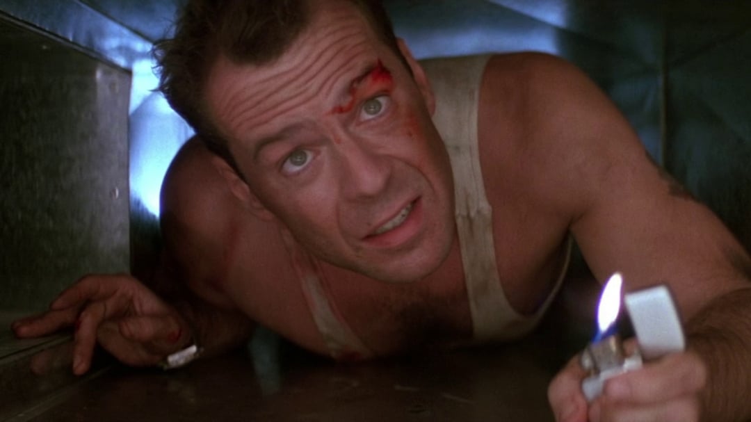 A man in a dirty tank top crawls through a narrow, metallic air duct, holding a lit lighter in front of him. He has a cut on his forehead and looks up with a concerned expression.