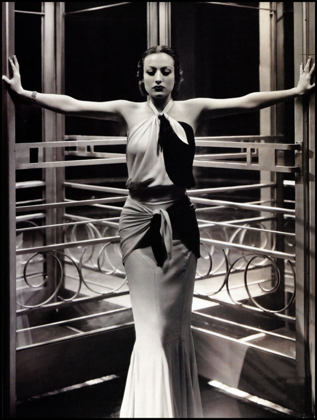 A woman in a glamorous, sleeveless evening gown stands confidently with arms outstretched, pressing against the sides of a revolving glass door adorned with geometric metal designs. The image is in black and white.