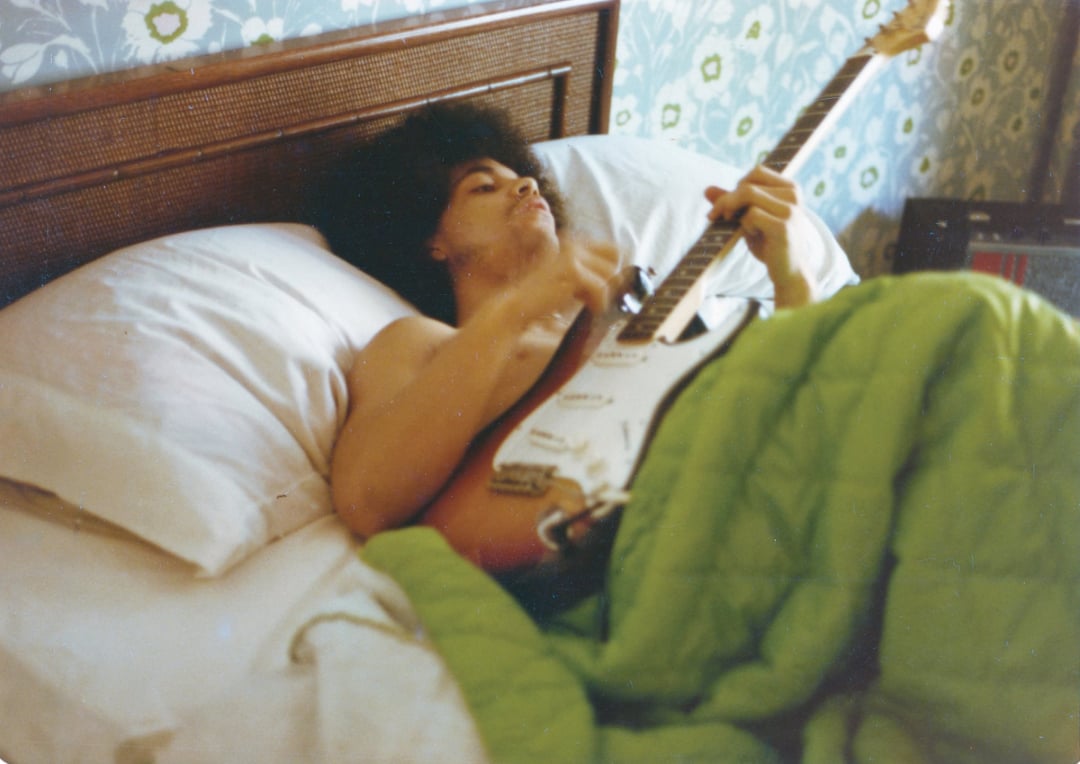 A person with curly hair lies in bed under a green blanket, shirtless, playing an electric guitar. The headboard is wooden, and the wall behind has a floral pattern.