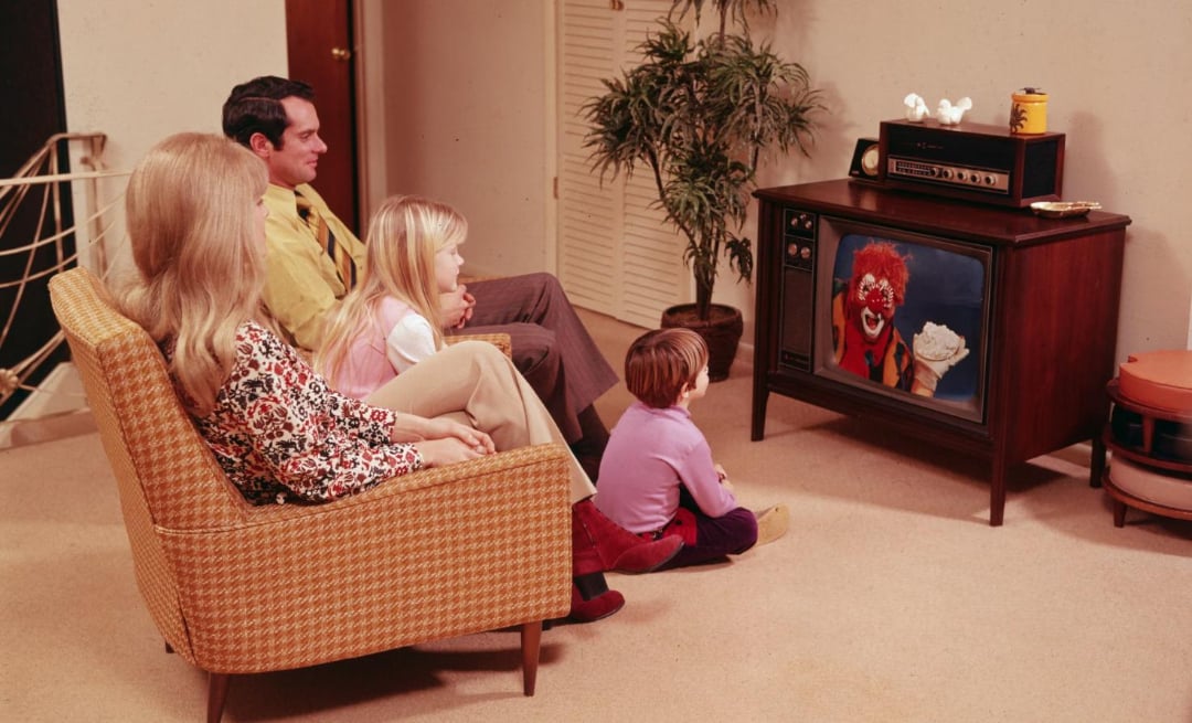 A family of four, two adults and two children, watches a television showing a clown holding a pie. The adults sit on a patterned couch, while the children sit on the floor in a cozy, vintage living room.