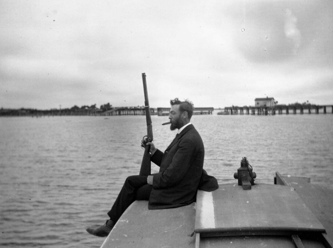 A man in a suit sits on the edge of a docked boat, holding a shotgun and looking out over the water. A pier and several buildings are visible in the background under a cloudy sky.