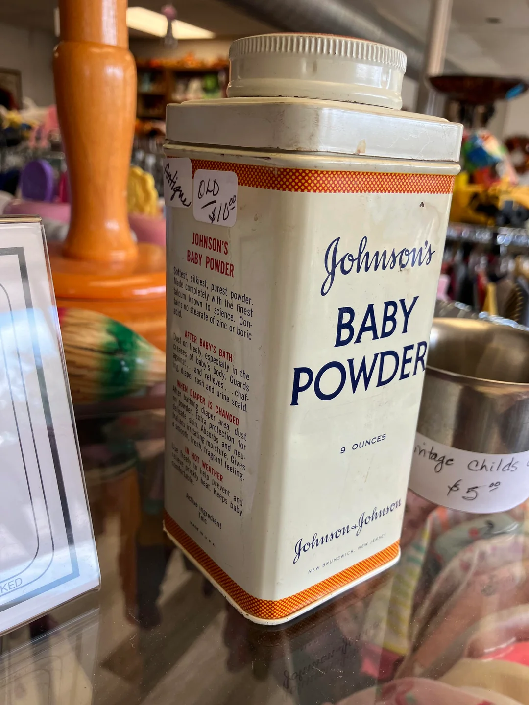 A vintage metal tin of Johnson’s Baby Powder sits on a glass display shelf in a shop, priced at $10.00. The container is white with orange and blue text and holds 9 ounces. Other items are visible in the background.