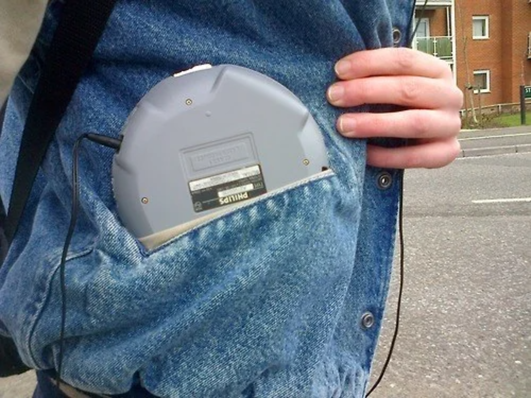 A person wearing a denim jacket puts a portable CD player into their large jacket pocket while standing near a street.