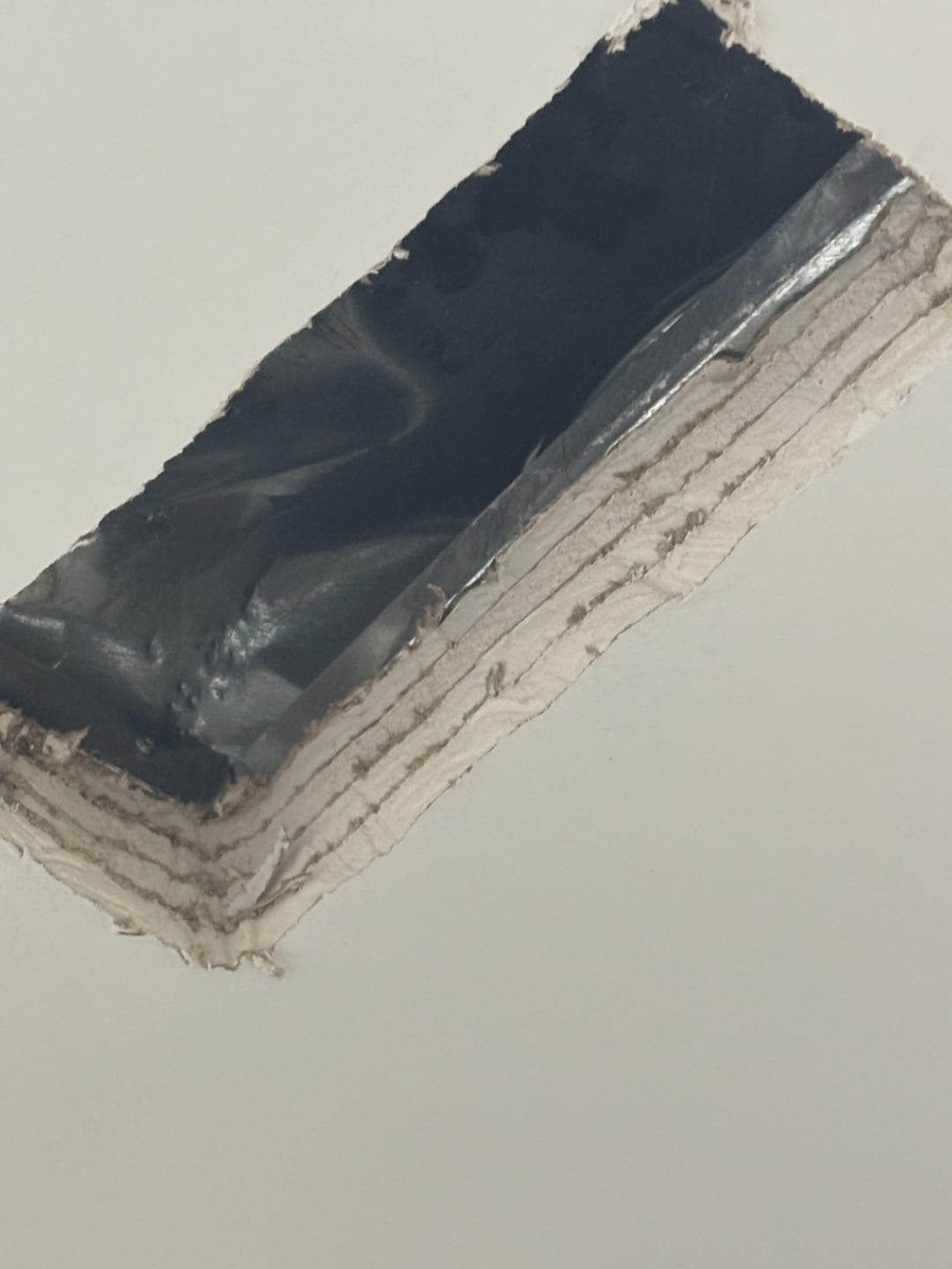 A close-up of a rectangular hole cut into a white wall or ceiling, revealing layers of drywall and a dark, shiny material inside the opening.