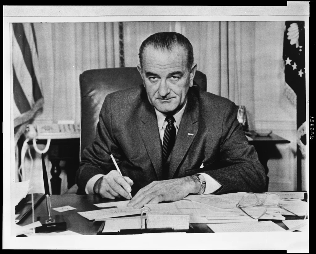 A man in a suit sits at a desk covered with papers, holding a pen and looking at the camera. An American flag and part of a presidential seal are visible in the background.