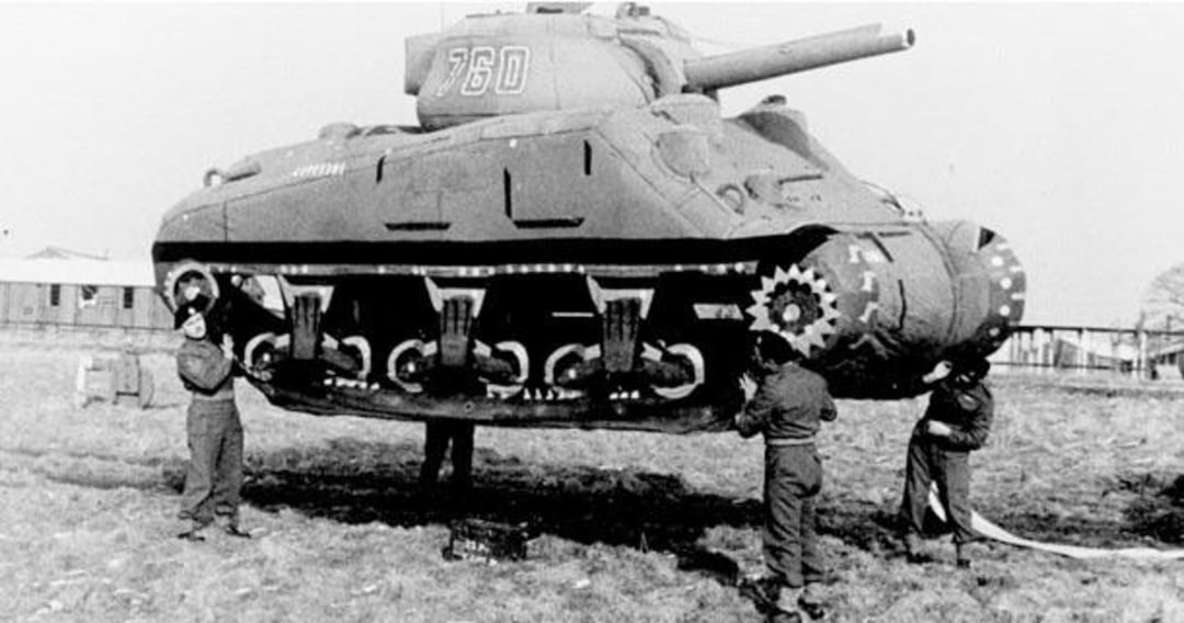 Three soldiers lift a large, lightweight tank prop, revealing it as a decoy. The tank appears to be floating above the ground, exposing its hollow underside. A building and trees are visible in the background.