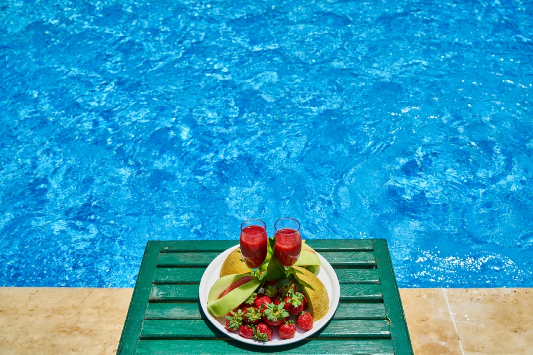 A plate with two glasses of red juice, sliced melon, and strawberries sits on a green table next to a bright blue swimming pool.
