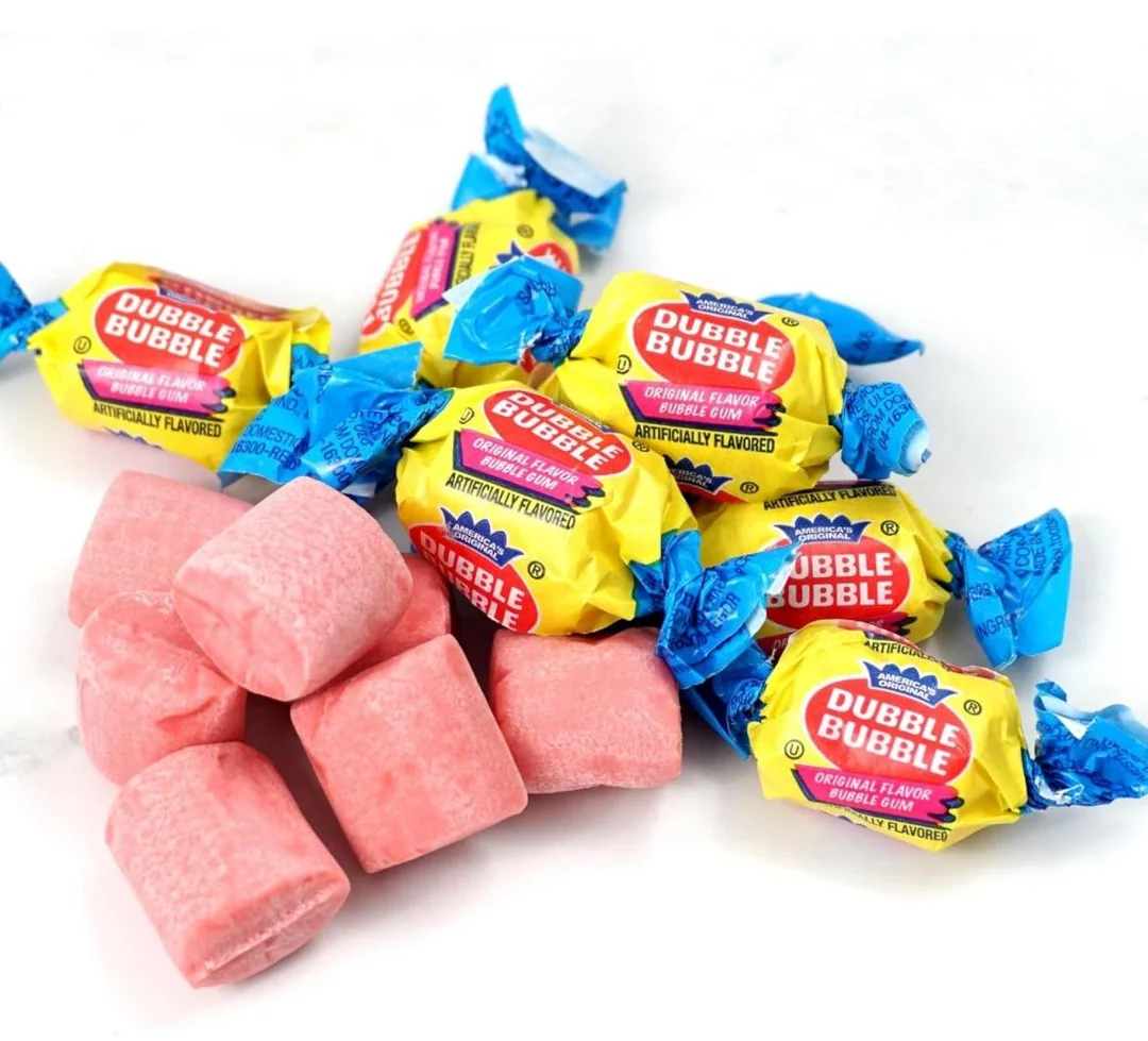 Several pieces of Dubble Bubble gum in yellow and blue wrappers sit next to unwrapped pink gum pieces on a white background.