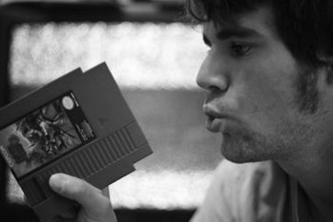 A person holds a retro video game cartridge and appears to be blowing on it, possibly to clean it. The image is in black and white.