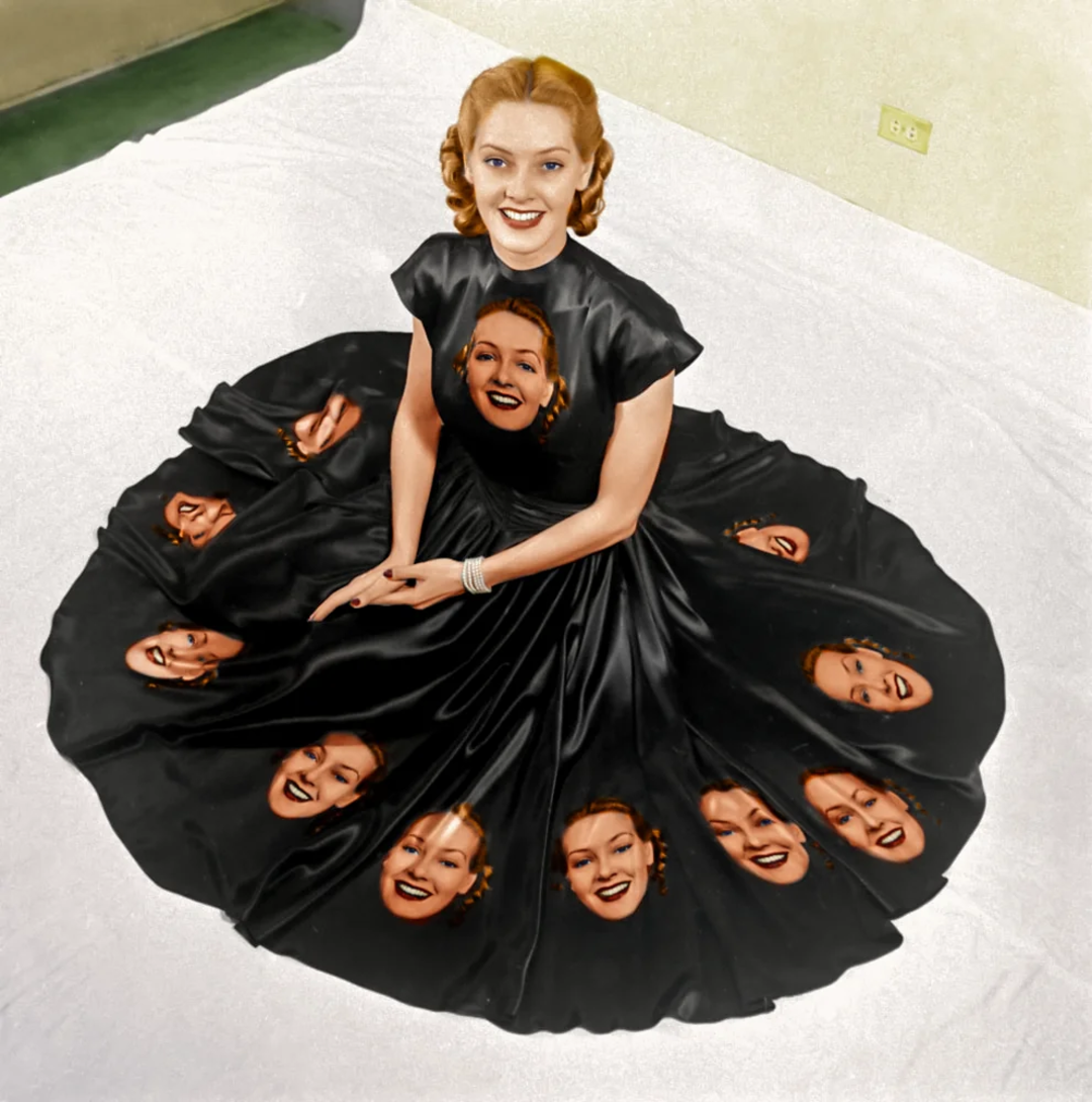 A smiling woman in a black dress sits on a white bed. The skirt of her dress is spread out in a circle and features multiple images of her own smiling face printed on the fabric.