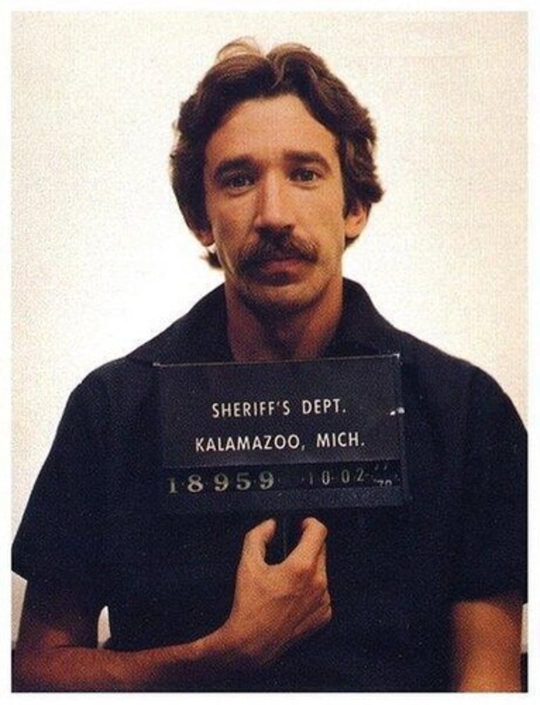 A man with short brown hair and a mustache holds a sign reading "Sheriff's Dept. Kalamazoo, Mich." in a mugshot-style photo against a plain background.