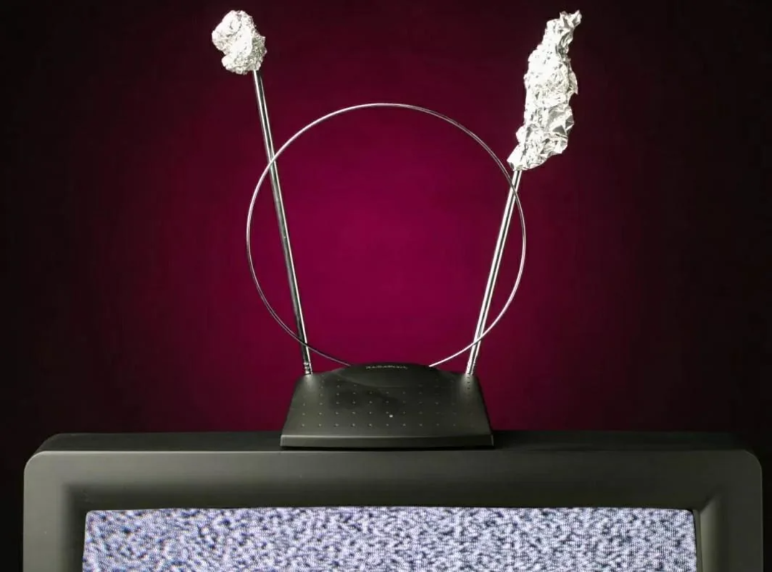 A TV antenna with aluminum foil wrapped around its tips sits on an old television displaying static, set against a dark red background.