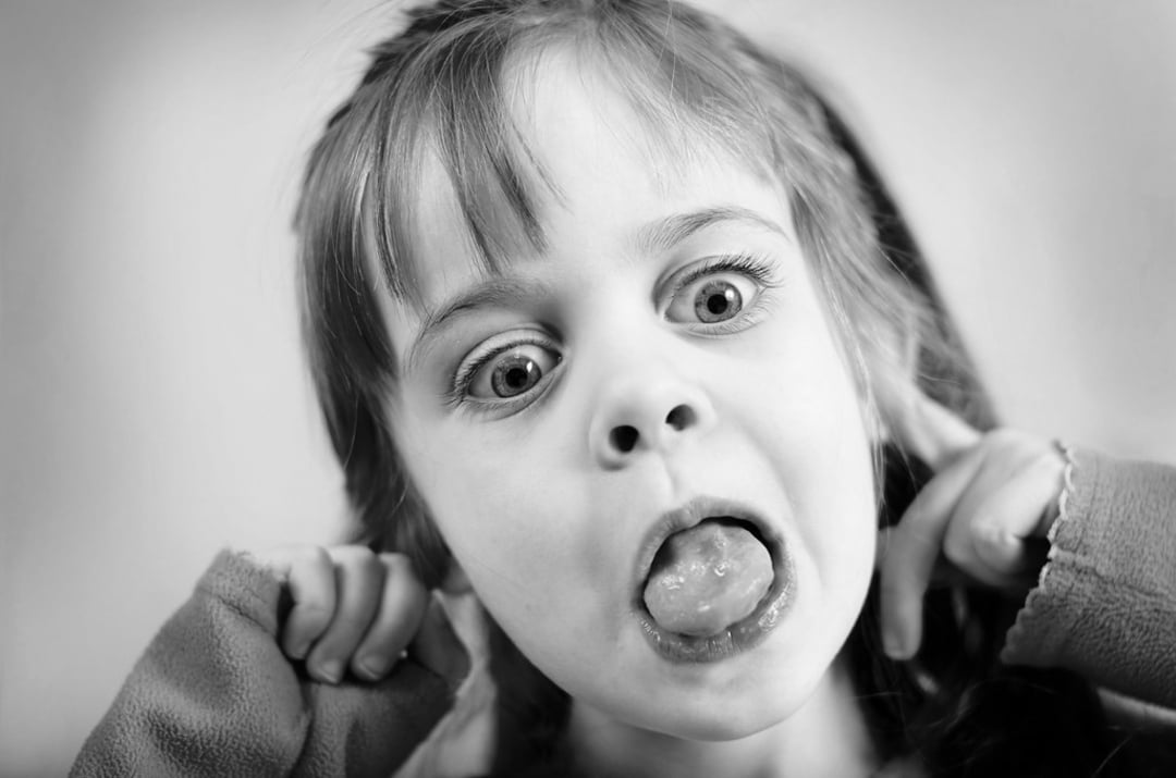 A young girl makes a funny face, sticking out her tongue, widening her eyes, and pulling at her ears with both hands. The image is in black and white.
