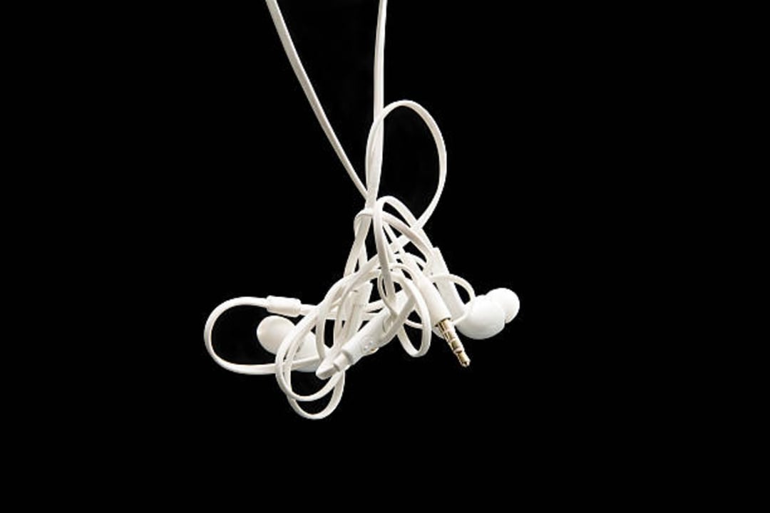 A pair of white wired earphones are tangled together, suspended against a solid black background. The 3.5mm audio jack and earbud tips are visible among the twisted cables.