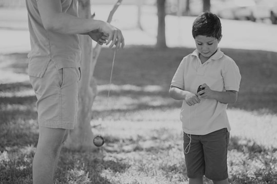 A man and a young boy stand on grass outdoors. The man holds a yo-yo, and the boy is looking down, winding the string around his hand. Trees and sunlight are visible in the background. The image is in black and white.