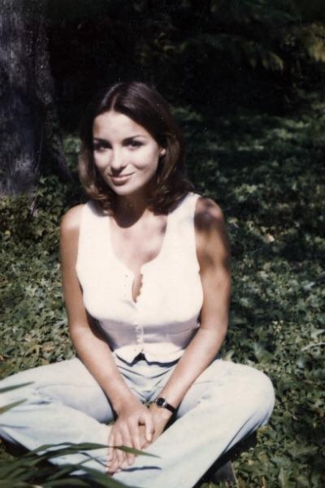 A young woman with shoulder-length brown hair sits cross-legged on grass, wearing a sleeveless white top and light blue jeans. She is outdoors, smiling softly, with greenery and sunlight in the background.