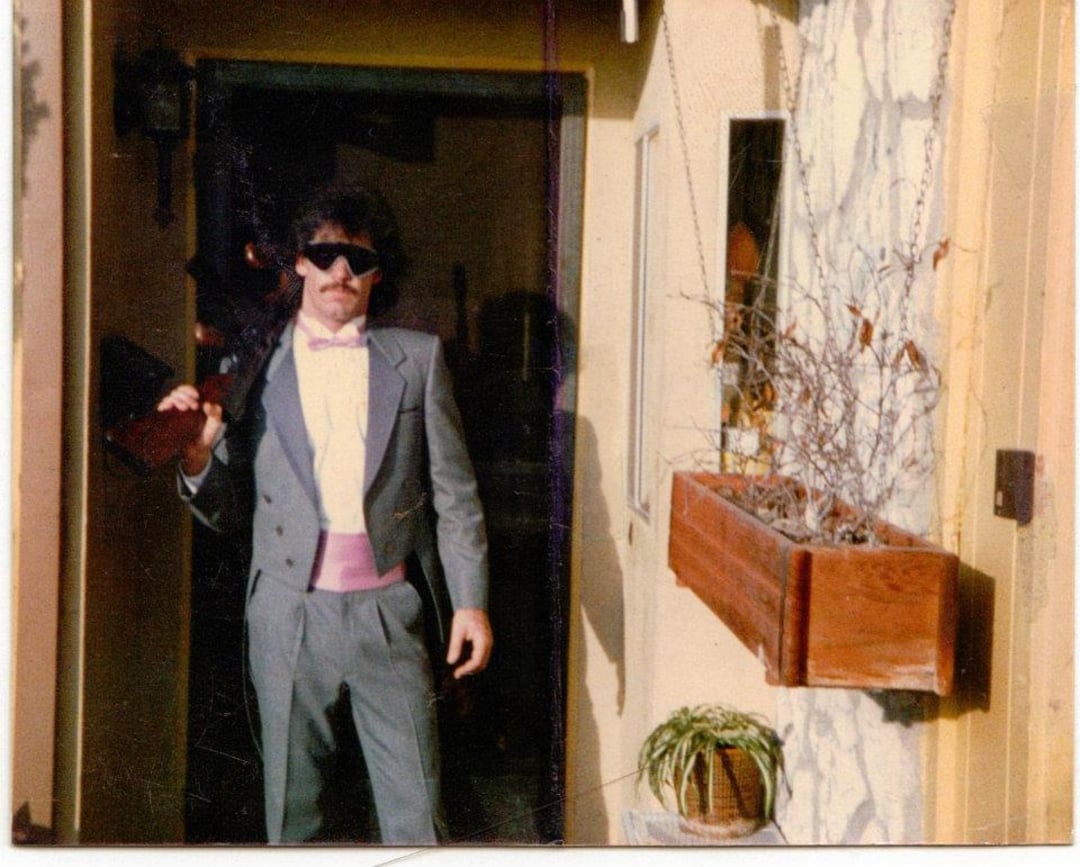 A man in a gray tuxedo with a pink cummerbund and bow tie stands outside a doorway, holding a jacket over his shoulder and wearing dark sunglasses. A planter with dry branches and a small potted plant are nearby.