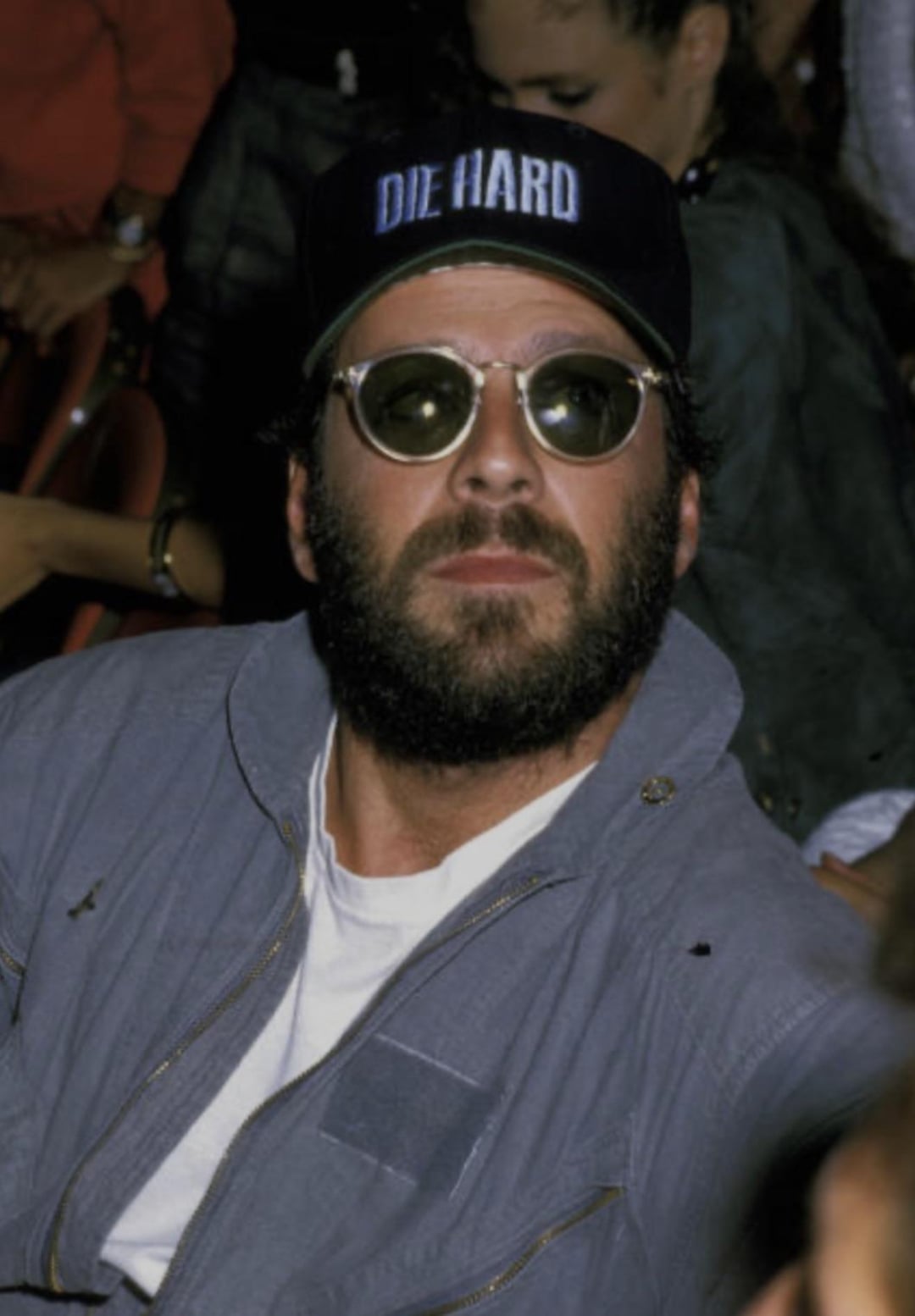 A man with a beard and sunglasses wears a black "DIE HARD" cap and a gray jacket while sitting in a crowd.