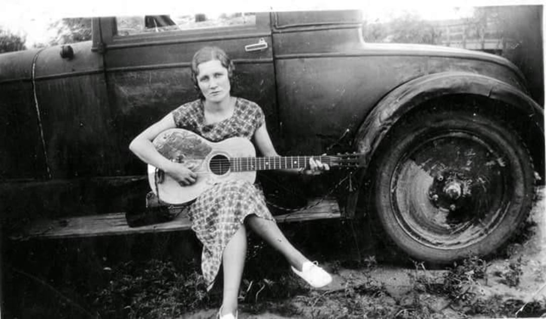 A woman in a patterned dress sits on the running board of an old car, playing an acoustic guitar. She looks at the camera with a calm expression. The background is outdoors, with some grass and foliage visible.