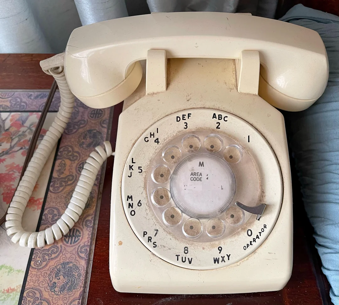 A beige rotary dial telephone with a coiled cord sits on a wooden surface next to a blue pillow and a colorful patterned mat.