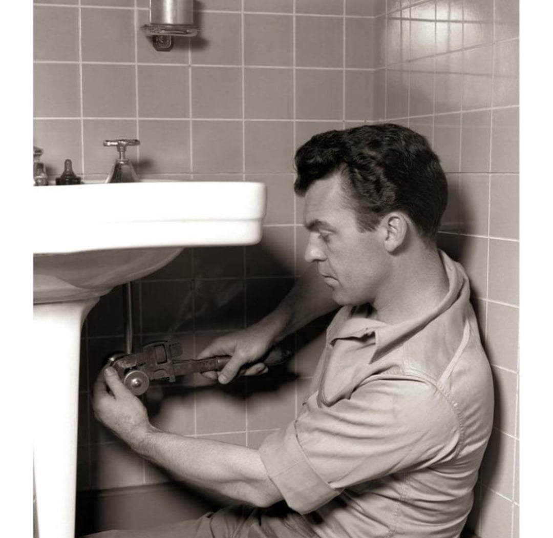 A man in work clothes is kneeling on a bathroom floor, using a large wrench to fix the pipes under a sink. The room has tiled walls and appears to be well-lit.