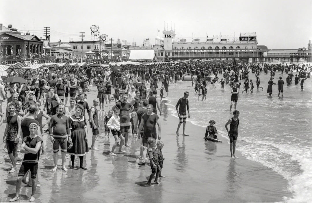 Crowds of people in vintage swimwear gather on a busy beach and in shallow ocean waves, with old-fashioned buildings and a pier visible in the background. The scene appears to be from the early 20th century.