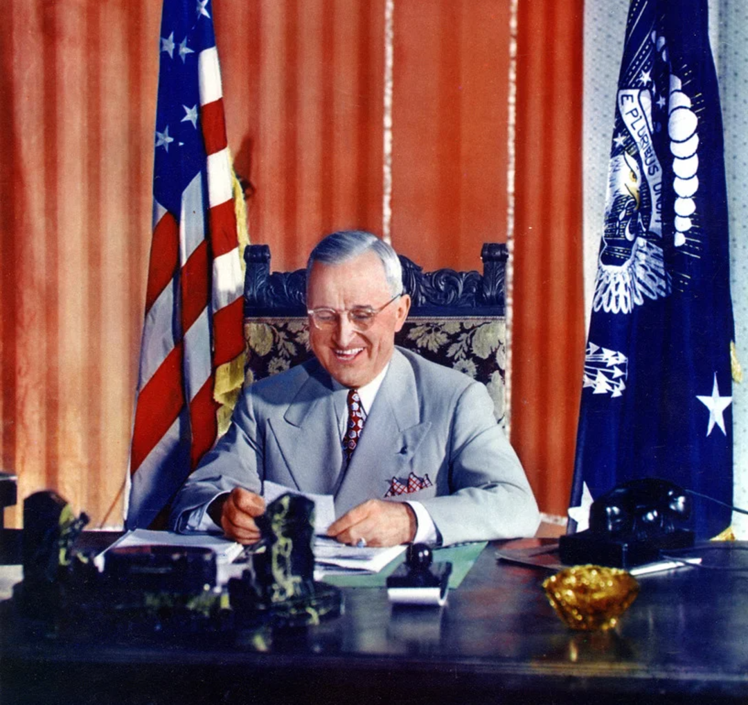 A smiling man in a light gray suit sits at a desk with papers, an American flag, and a presidential flag behind him, in an office with orange curtains.
